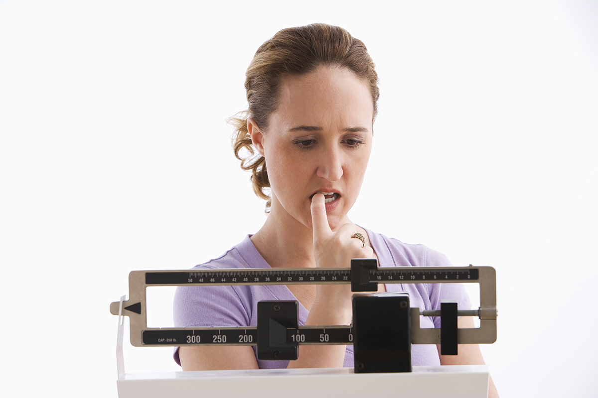 A young woman is standing on her scale and checking her weight. She has a worried expression on her face. Horizontal shot. Isolated on white.